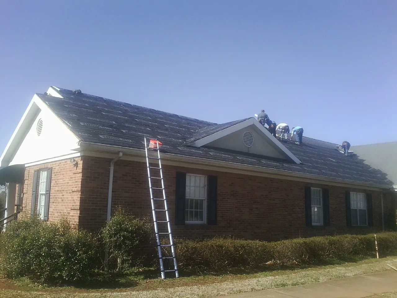 Crew tearing shingles off a brick ranch roof with a ladder set — Metal Roof Repair work by our crew in Merlin