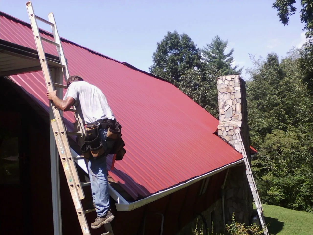 Roofer on a ladder beside a red metal roof and stone chimney — Soffit & Fascia Repair by Roofing Brothers in Merlin